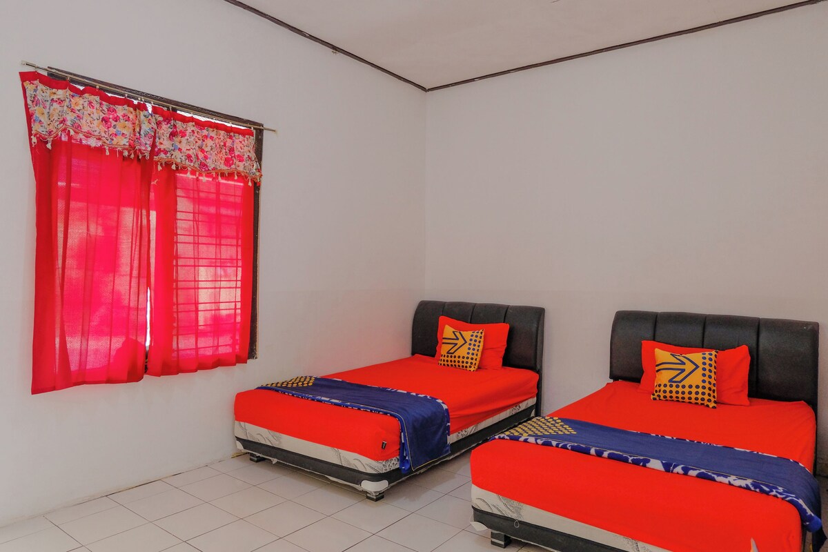 A room with two beds, each with red bedding and patterned pillows, a window with red curtains, and a tiled floor.