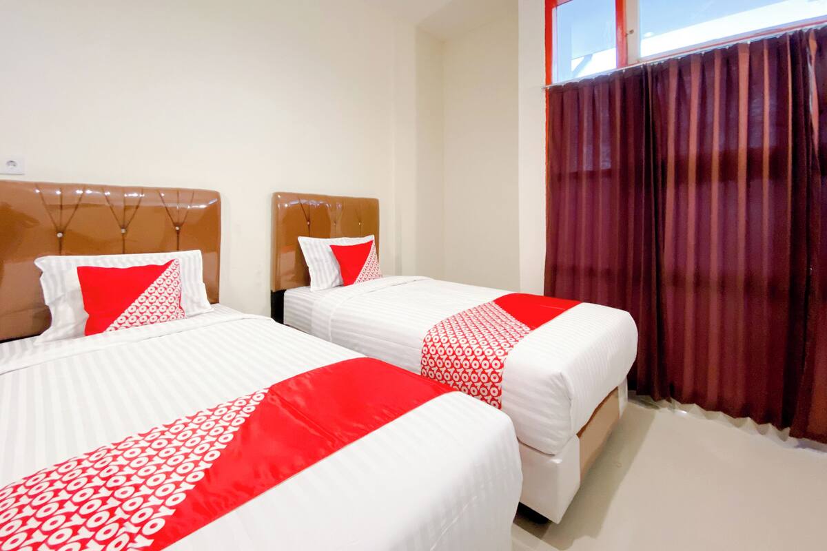 A hotel room with two beds, each with white bedding and red accents, a wooden headboard, and a window with red curtains.
