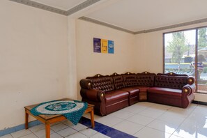 A living room with a brown leather sofa, a small wooden table with a patterned tablecloth, and a tiled floor.