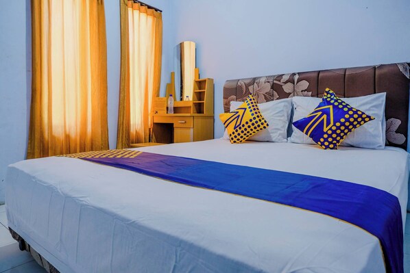 A neatly made bed with a blue and white bedspread, two pillows with geometric patterns, a wooden nightstand with a mirror, and a large window with curtains.