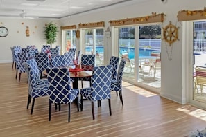 A room with a pool view, featuring a dining area with patterned chairs and a table, wooden flooring, and a large window.