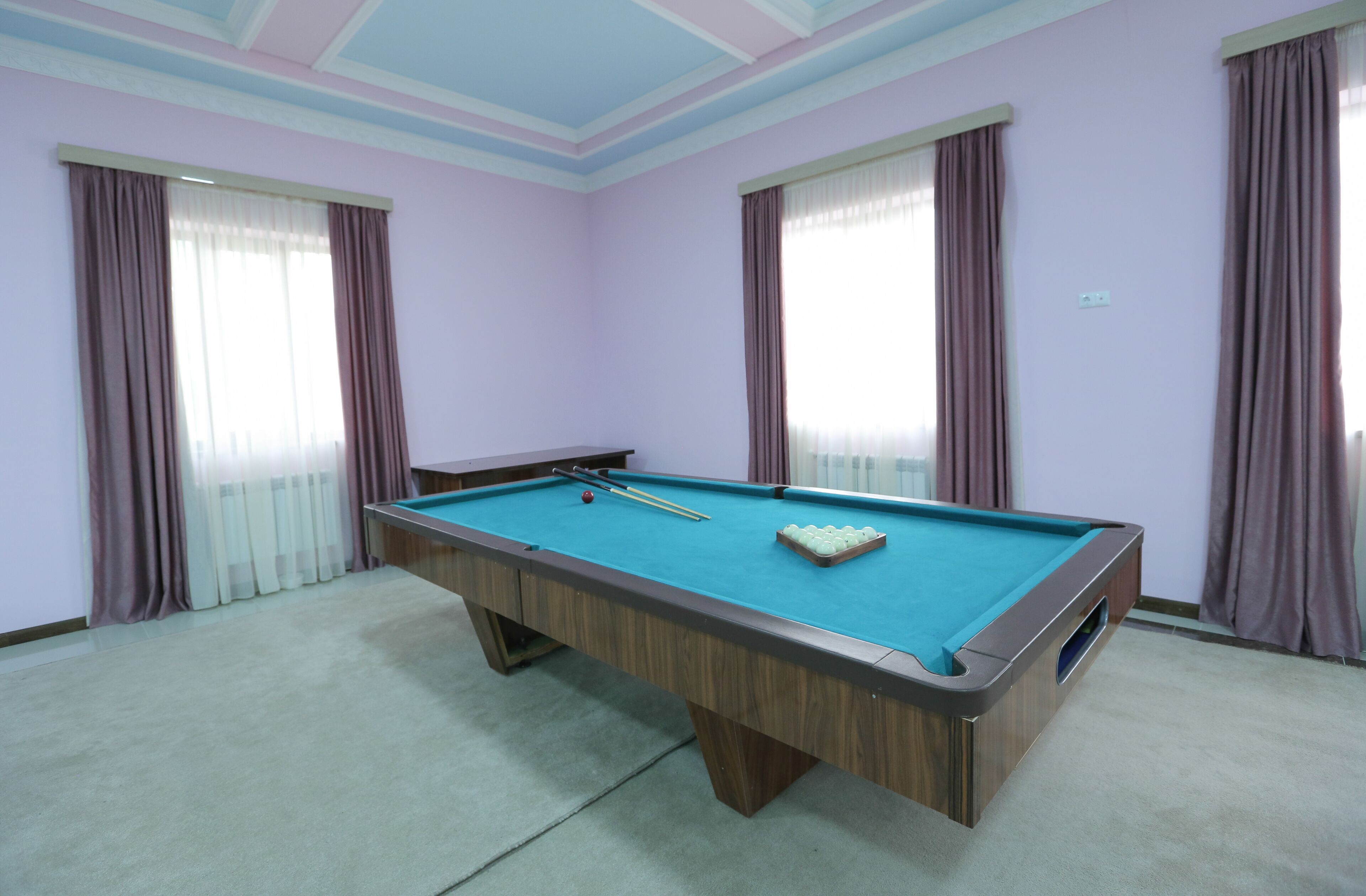 A pool table in a room with purple walls and large windows with curtains.