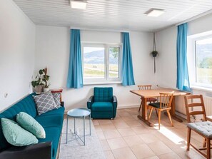 Een kamer met een blauwgroene bank, een klein rond tafeltje, een houten eettafel met stoelen en uitzicht op het omliggende landschap door een raam.