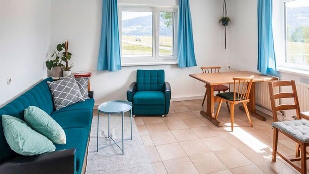 Ein Zimmer mit einem türkisfarbenen Sofa, einem kleinen runden Tisch, einem Holzesstisch mit Stühlen und Blick ins Freie durch ein Fenster.