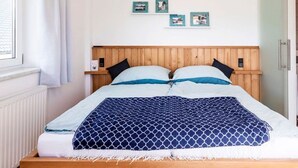 Ein Schlafzimmer mit Holz-Kopfteil, einem Bett mit blau-weißer Bettwäsche, einem Fenster mit weißen Vorhängen und drei gerahmten Bildern an der Wand.