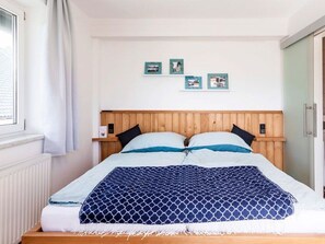 Un dormitorio con cabecera de madera, cama con ropa de cama azul y blanca, ventana con cortinas blancas y tres cuadros en la pared.