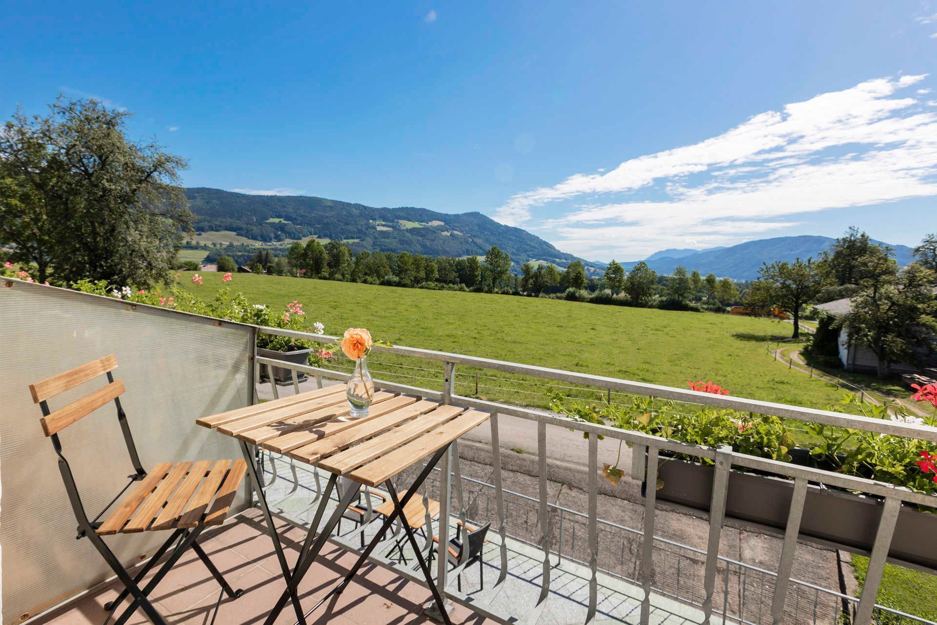 Ein Balkon mit Holztisch und -stühlen, mit Blick auf ein grünes Feld und Berge.