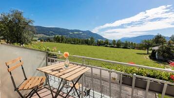 Ein Balkon mit Holztisch und -stühlen, mit Blick auf ein grünes Feld und Berge.