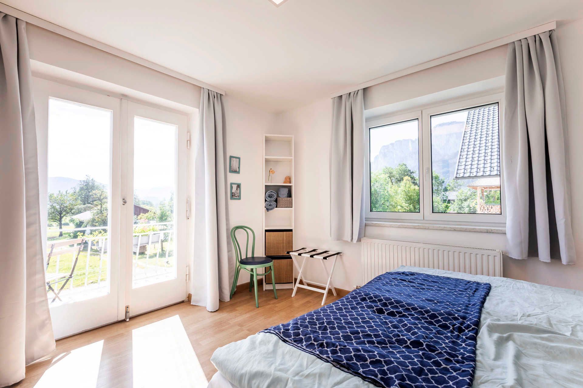 Ein Schlafzimmer mit einem Bett, einem Sessel, einem Heizkörper, einem Fenster mit Blick auf Grünflächen und einer Tür zu einem Außenbereich.
