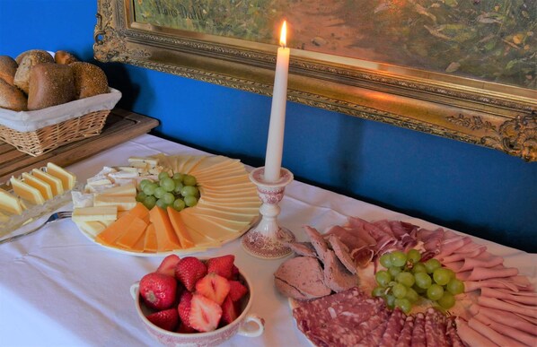 A table with cheese, grapes, strawberries, and various meats, a lit candle, and a framed painting on a blue wall.