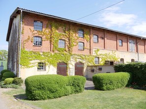 Un bâtiment de deux étages en briques, doté de nombreuses fenêtres, d’une porte centrale et d’un petit jardin agrémenté de haies.