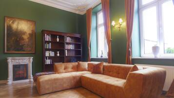 Ein Zimmer mit Ecksofa, Kamin, Bücherregal und einem großen Fenster mit Vorhängen.