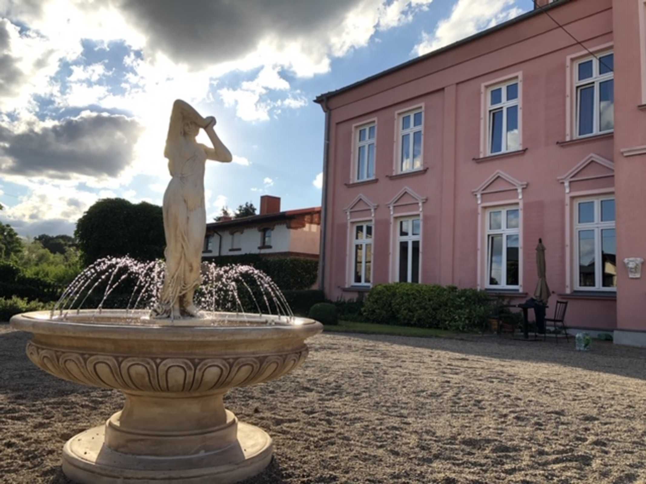 Eine klassische Statue auf einem Brunnen vor einem rosafarbenen Gebäude mit zahlreichen Fenstern.