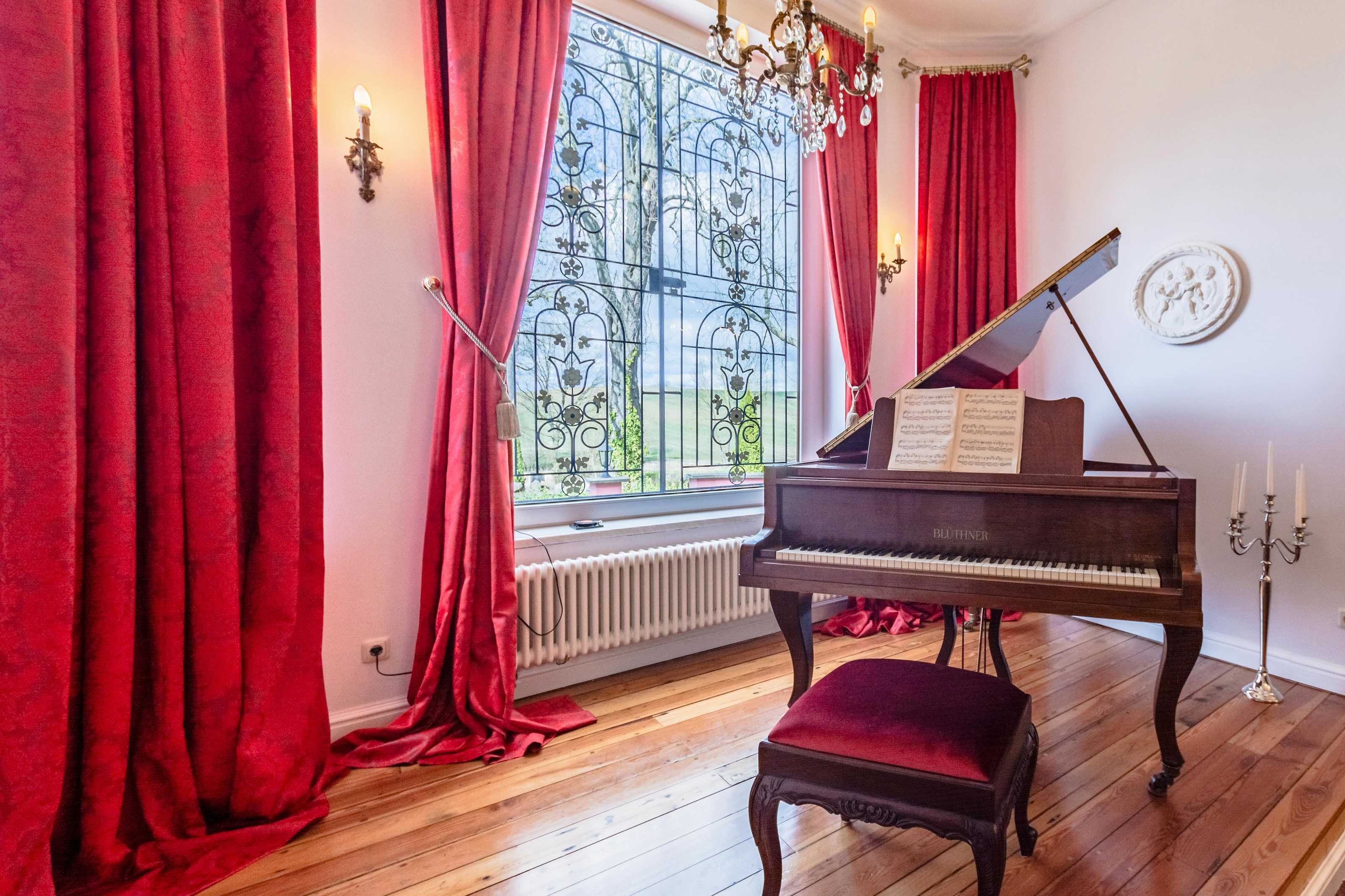 A grand piano, red curtains, a radiator, and a chandelier in a room with wooden flooring.