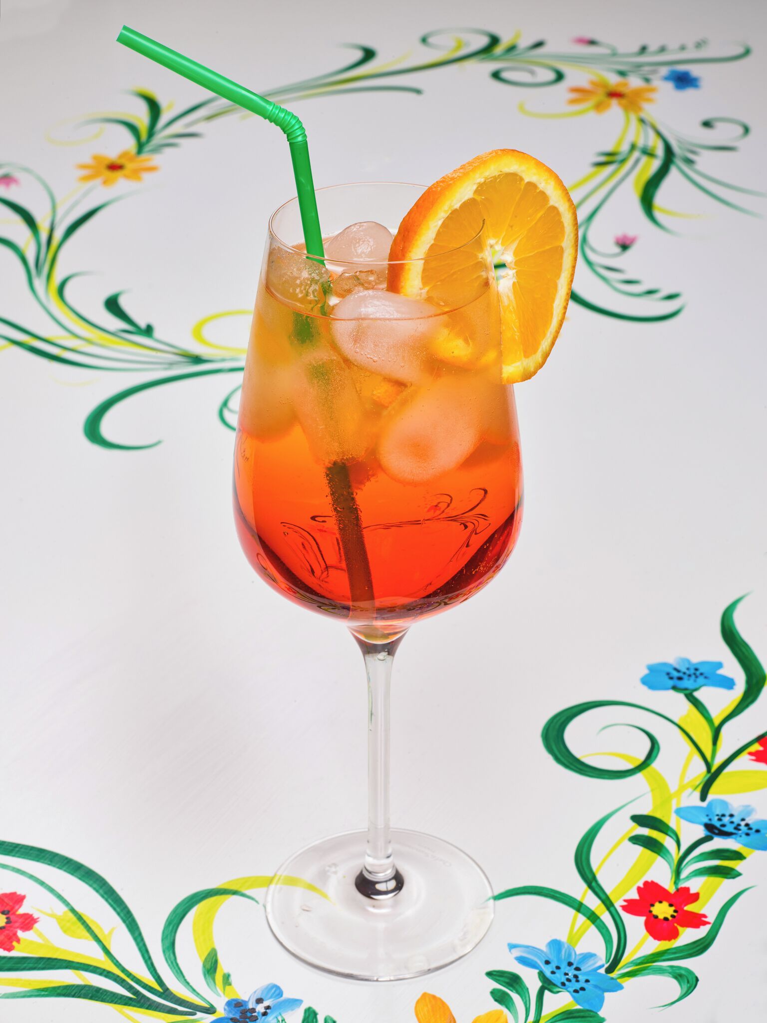 A glass of red beverage with ice cubes, a slice of orange, and a green straw.