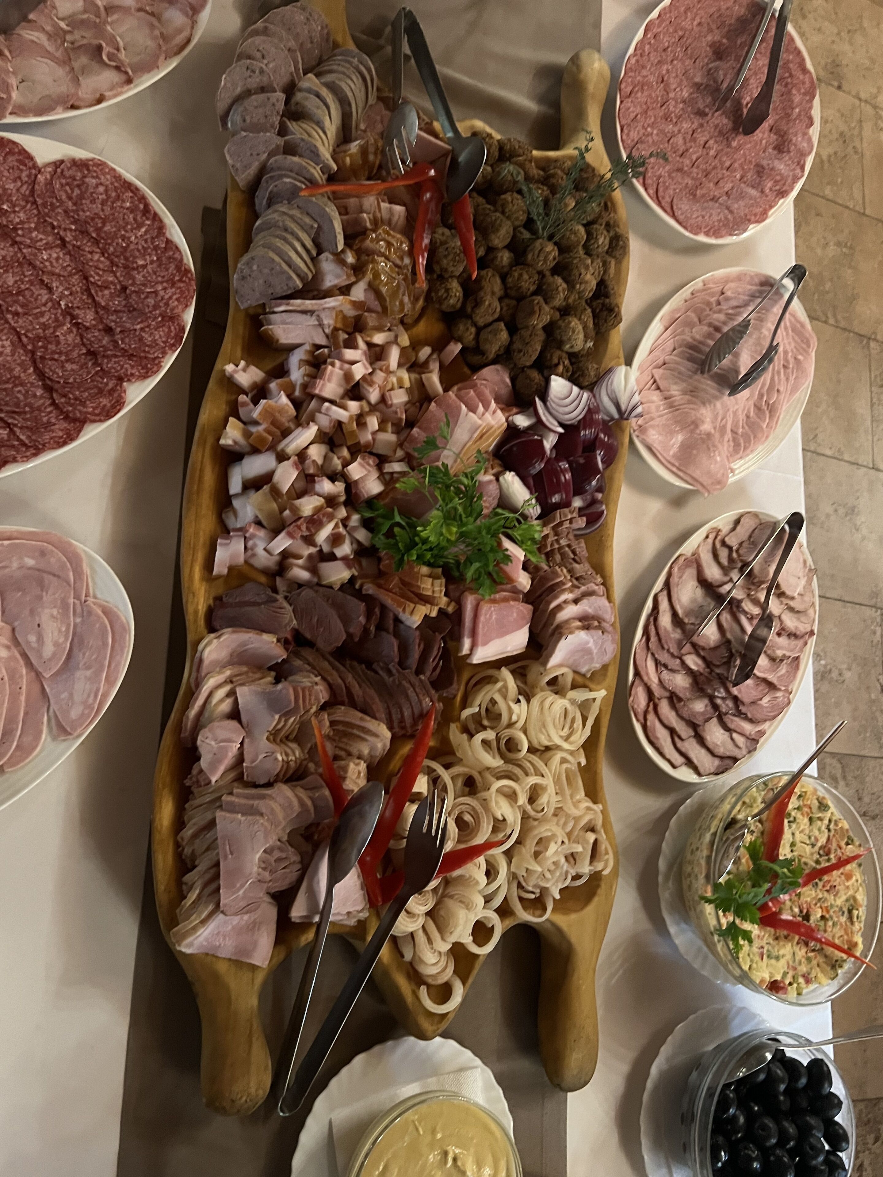 A variety of meats and cheeses arranged on a wooden board, with tongs and a sprig of parsley for decoration.