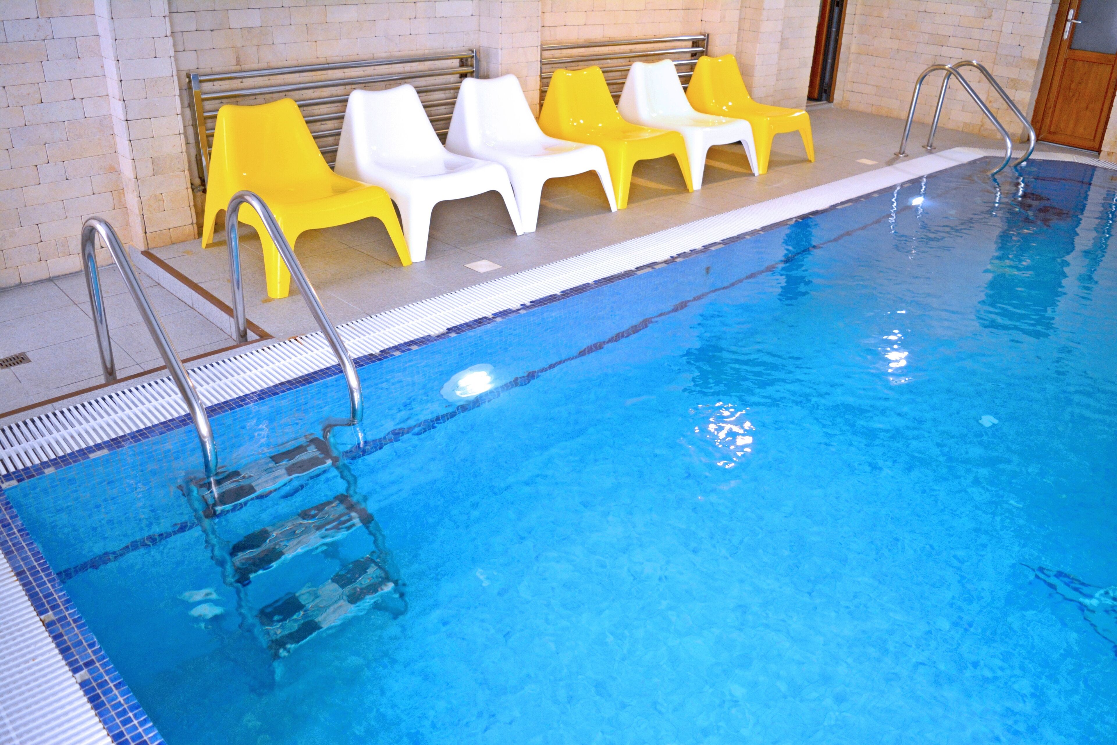 Une piscine intérieure à l’eau d’un bleu limpide, avec un escalier en métal et des chaises longues jaunes et blanches disposées le long du bord.