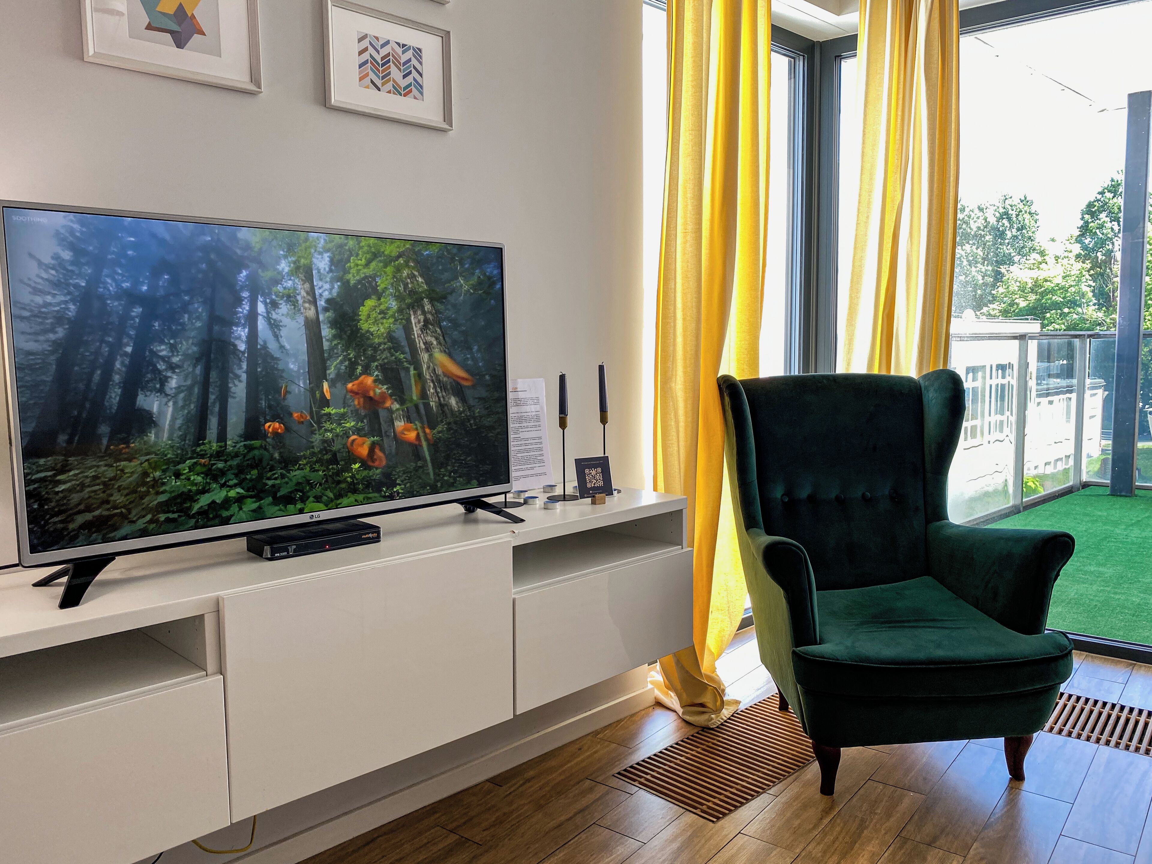 Un salon avec un téléviseur à écran plat, un fauteuil vert foncé et une fenêtre ornée de rideaux jaunes.