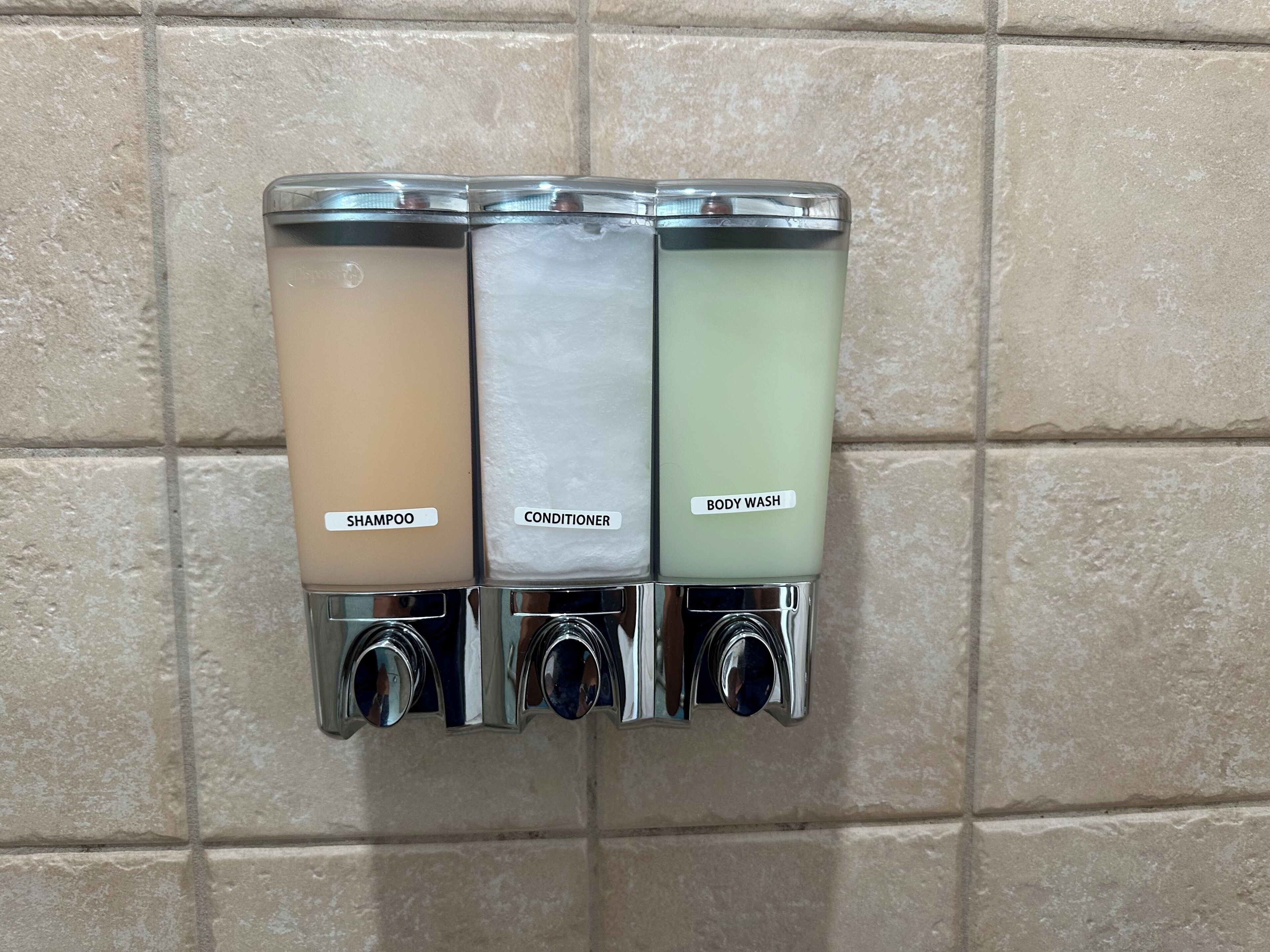 A wall-mounted dispenser with three compartments labeled "SHAMPOO," "CONDITIONER," and "BODY WASH."