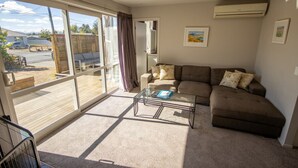 Ein geräumiger Wohnbereich mit einer großen Schiebeglastür, die auf eine Terrasse im Freien führt, ein braunes Sofa mit floralen Kissen, ein Glastisch und eine Klimaanlage an der Wand.