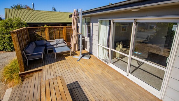 Eine Holzterrasse mit einer Couch und einem Tisch, eine Glastür und ein grünes Dach.
