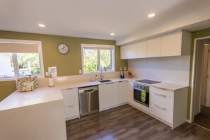 A modern kitchen with white cabinets, a dishwasher, an oven, and a sink. There are two windows providing natural light and a clock on the wall.