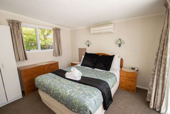 A bedroom with a bed, two bedside tables, a dresser, an air conditioning unit, and a window with curtains.