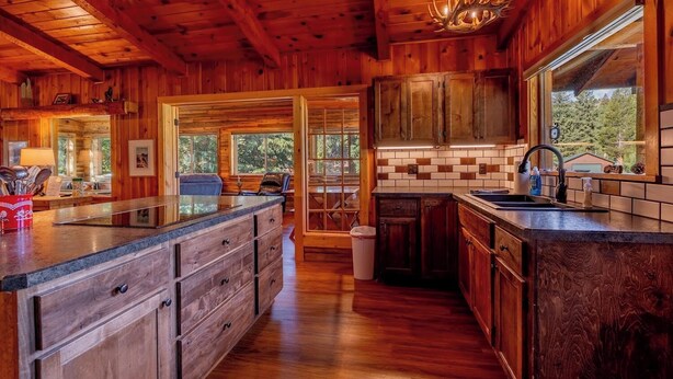 Interior de una cabaña de madera con cocina, que incluye una isla grande, armarios de madera y vistas a través de las ventanas.