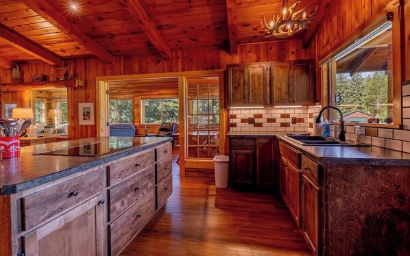 Interior de una cabaña de madera con cocina, que incluye una isla grande, armarios de madera y vistas a través de las ventanas.