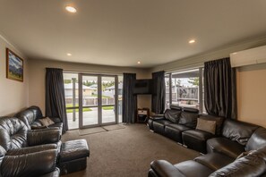 Ein geräumiges Wohnzimmer mit einem großen Schiebeglasfenster, das in einen Garten führt, Ledersofas, einem wandmontierten Flachbildfernseher und einem kleinen Tisch.
