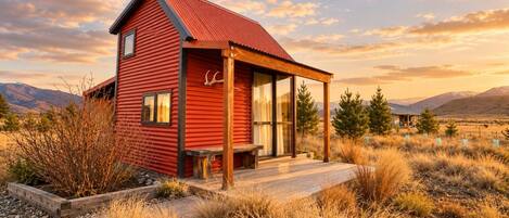 A red cabin with a porch, surrounded by dry grass and mountains in the background.