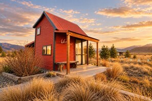 A red cabin with a porch, surrounded by dry grass and mountains in the background.
