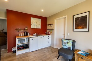 Cocina compacta con microondas, horno y armarios blancos, una encimera de madera con utensilios de cocina, una silla con cojín colorido y un cuadro en la pared.