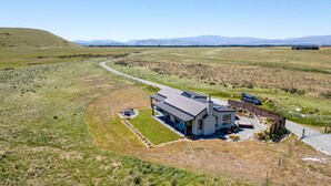 A modern house with a large patio area, surrounded by open fields and a winding road.