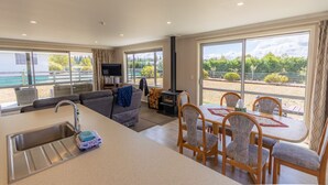 A modern living room with a sofa, dining table, and chairs, a kitchen area with a sink, and large windows offering a view of the outdoors. - New with a View, Twizel - FREE WIFI (Twizel)