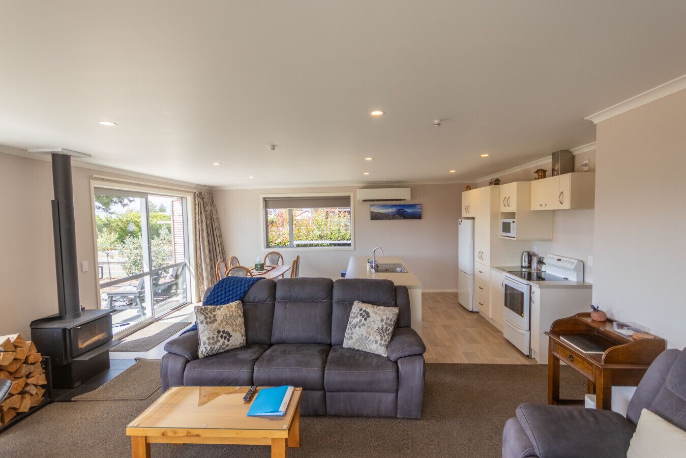 A modern living room with a grey sofa, a wooden coffee table, and a kitchen area with white cabinets and appliances.
