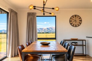 A dining area with a wooden table, chairs, a large window with a mountain view, a chandelier, and a wall clock.