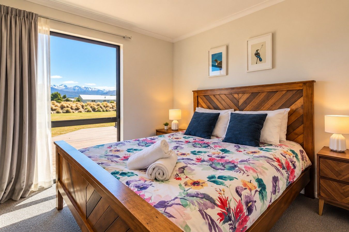 A hotel room with a large bed, a wooden headboard, two bedside lamps, a window with a view of mountains, and a door with curtains.