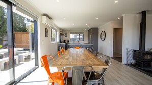 Un comedor amplio con una larga mesa de madera y sillas modernas, una cocina al fondo y un gran ventanal con vistas al exterior.