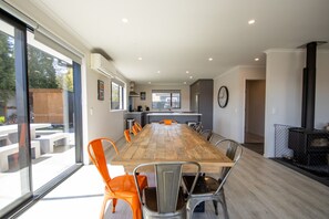 A spacious dining area with a long wooden table and modern chairs, a kitchen in the background, and a large window offering a view of the outdoors.