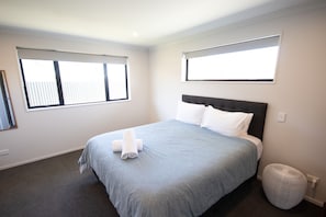 A bedroom with a bed, two windows, a wall-mounted outlet, and a round ottoman.