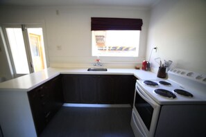 A small kitchen with a white stove, dark cabinets, a window with a brown valance, and a sink.