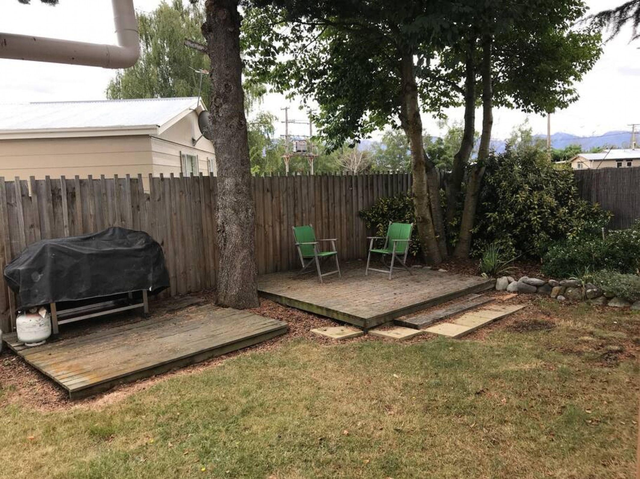 Un jardin avec une terrasse en bois, deux chaises vertes et un barbecue recouvert d’un tissu noir.