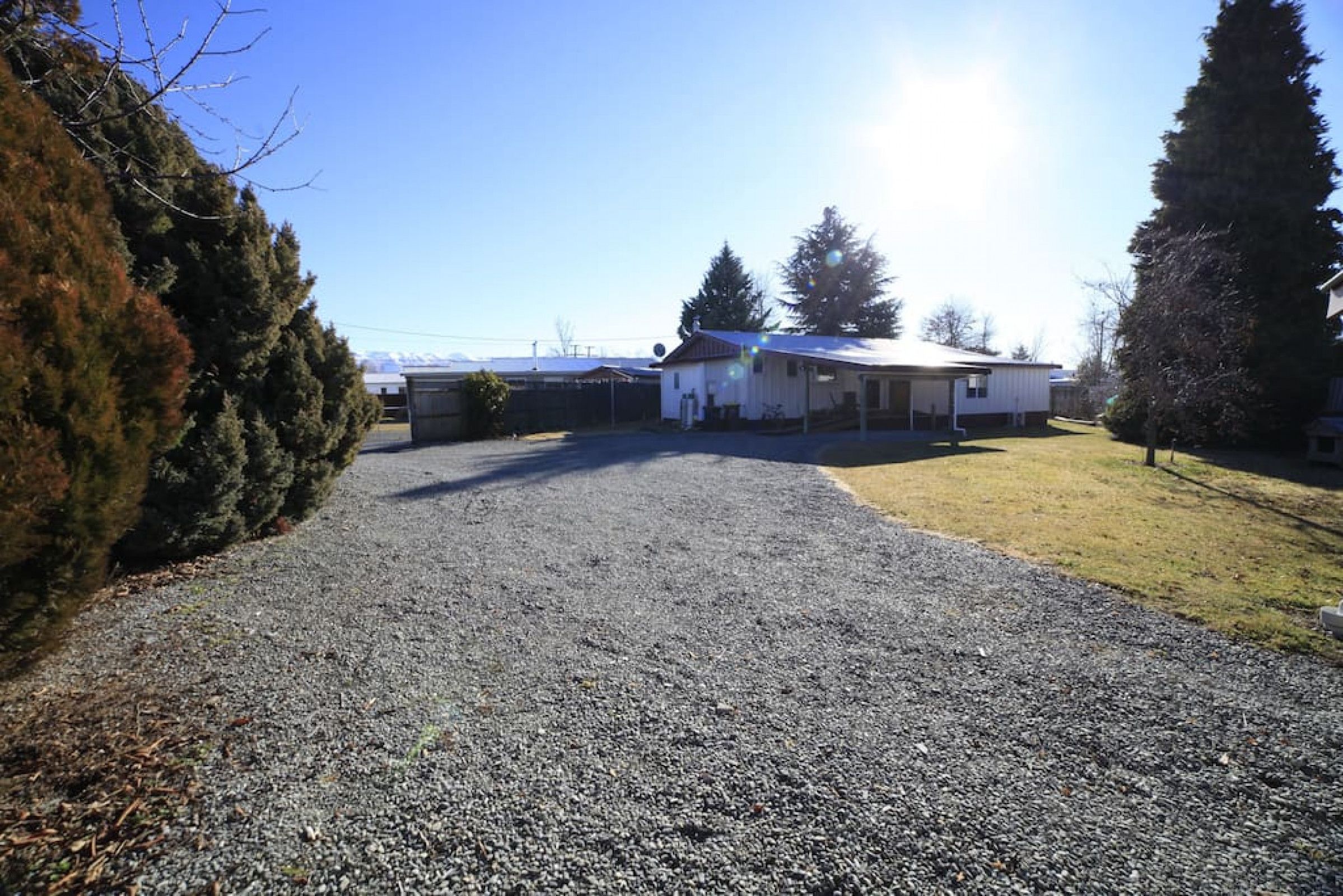 A gravel driveway leading to a white building with a covered porch, surrounded by trees and a clear sky.