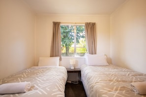 A twin room with two beds, a window with curtains, a bedside table with a lamp, and a view of greenery outside. - Holiday Haven - FREE WIFI (Twizel)