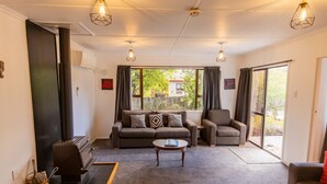 A cozy living room with a sofa, armchair, and a coffee table. There is a fireplace with a chimney, and a window with curtains. - Holiday Haven - FREE WIFI (Twizel)
