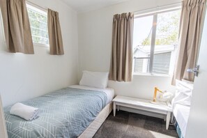 Een kleine kamer met een enkel bed, een raam, een nachtkastje met een lamp en een opgerolde handdoek op het bed.
