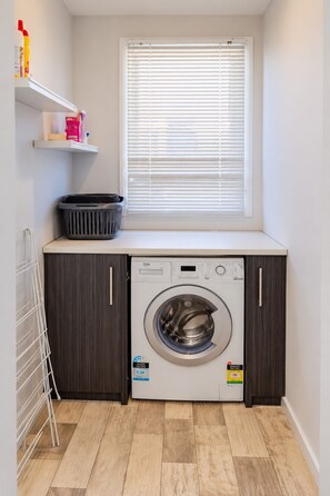 Une petite buanderie équipée d’un lave-linge à chargement frontal, d’un étendoir et d’une fenêtre avec des stores.