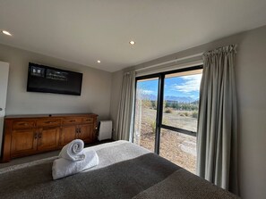 A hotel room with a large bed, a wooden dresser, a flat-screen TV, and a view of a mountainous landscape through a sliding glass door. - Big Sky, Big View - Family friendly and close to Mt Cook, FREE WIFI (Twizel)