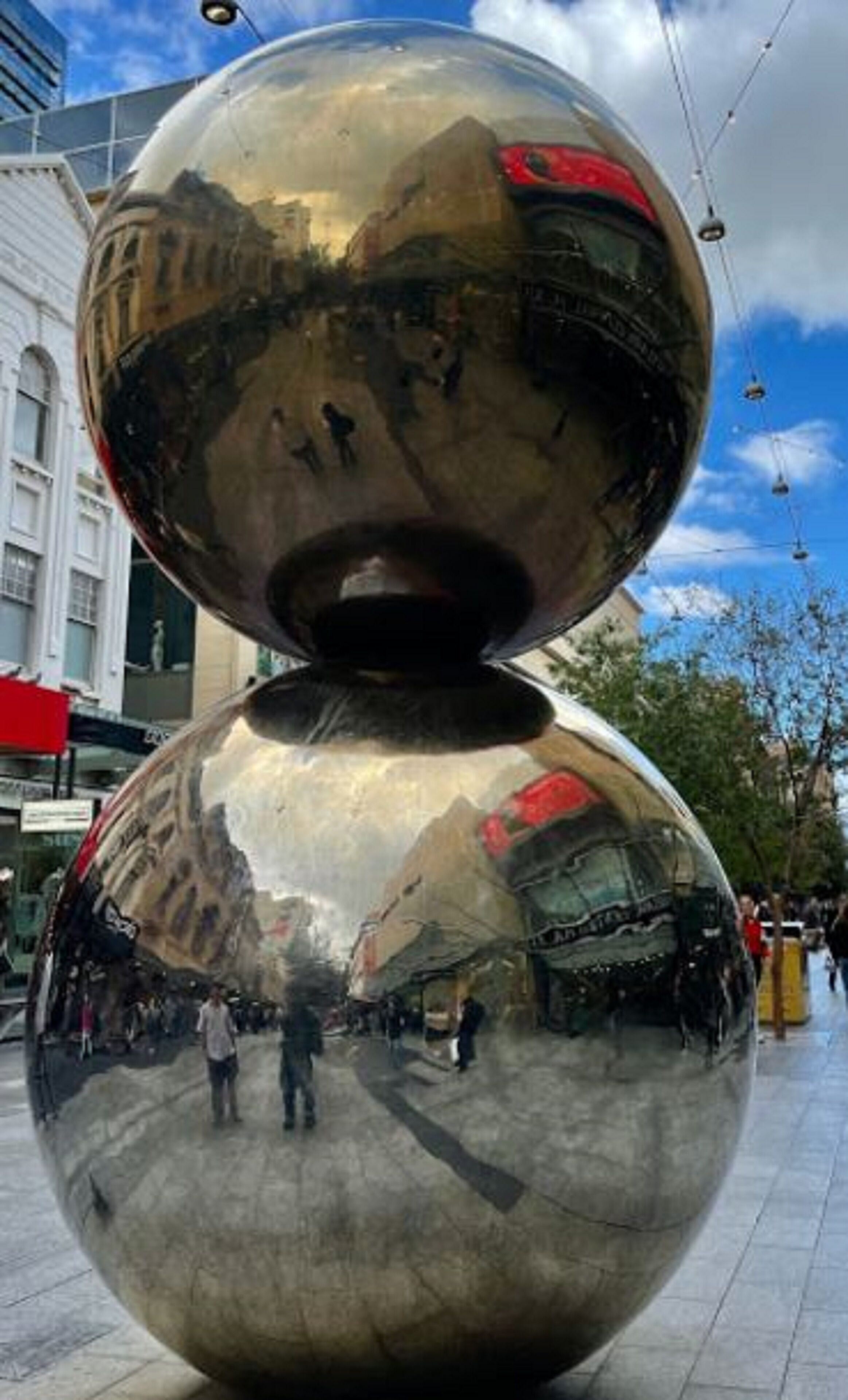 A spherical sculpture with a reflective surface, located in an urban setting with buildings and pedestrians in the background.